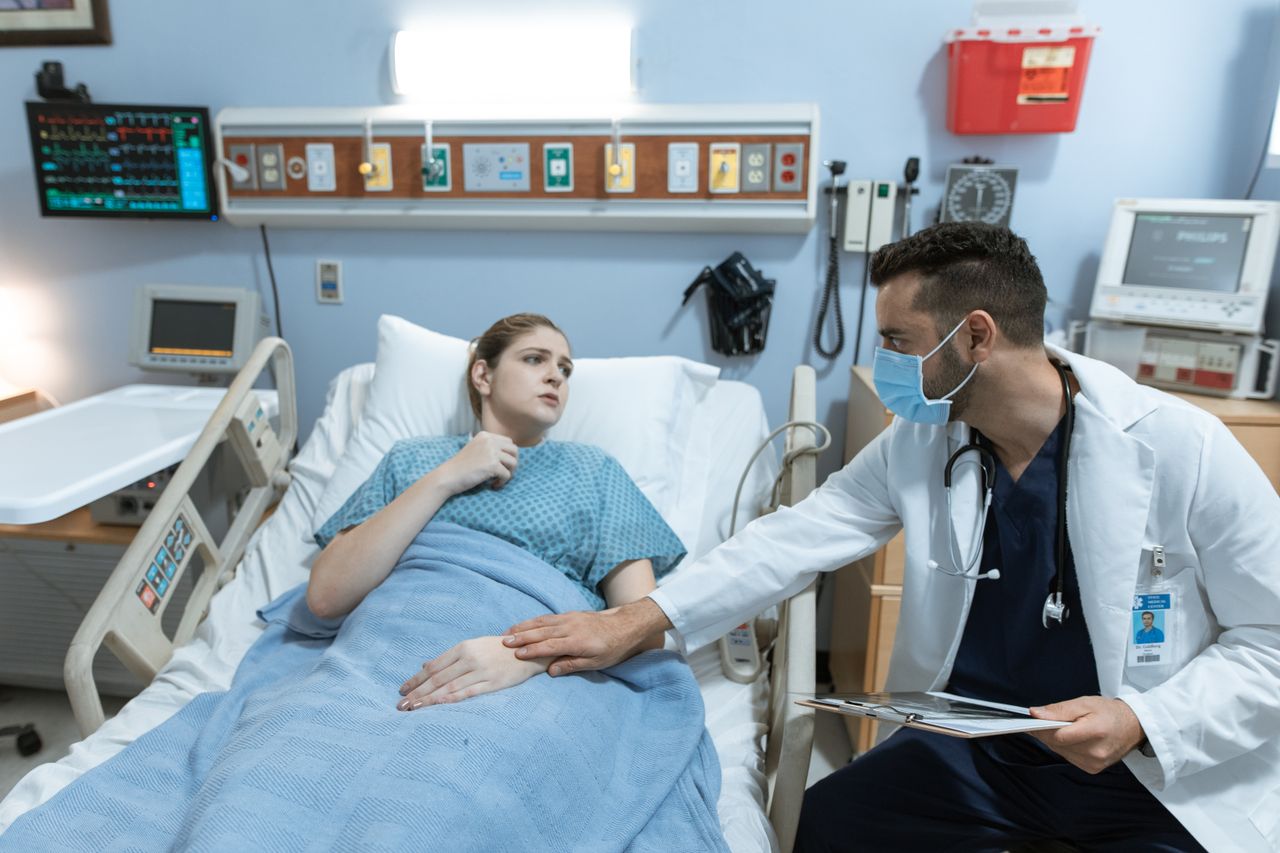 Male doctor with face mask comforts female patient in hospital bed with medical equipment.