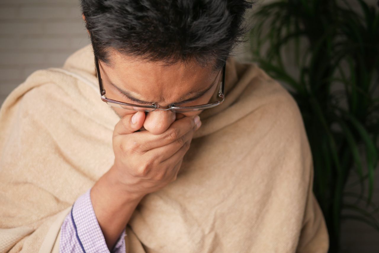 Adult wearing glasses coughs into hand indoors, indicating illness or flu symptoms.